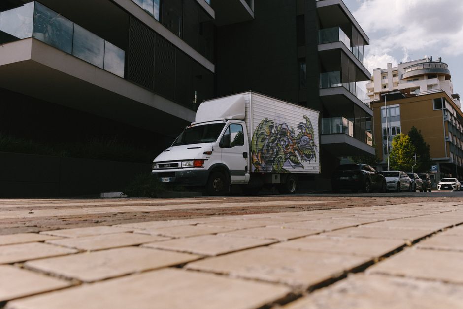 Two professional movers from Man with Van Woodlands are indoors during a home relocation process, carrying cardboard boxes. The man on the left, with curly hair and a beard, holds two stacked medium-s