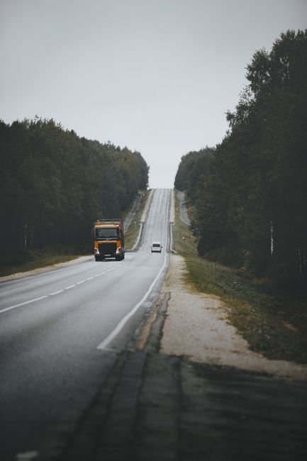 A straight rural road with two lanes, bordered by tall, dense green trees on both sides, leading into the distance under an overcast sky. A large black and orange moving truck is traveling away from the camera on the left side of the road, while a small white car is driving towards the camera along the right lane. The roadside includes a narrow dirt verge and grass strip, with slight elevation changes visible on the horizon. The scene captures the quiet and open atmosphere typical of regional areas, relevant to the context of house removals and home relocation logistics facilitated by Man with Van Woodlands.