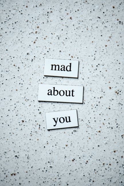 The image shows three small rectangular tiles arranged vertically on a speckled, light-colored surface. The tiles are white with black text, reading 'mad', 'about', and 'you' from top to bottom. They appear to be made of plastic or wood and are placed neatly with slight spacing between each. The background surface is textured, resembling a terrazzo or speckled countertop, with scattered small dark and light particles. This arrangement of tiles resembles a phrase, potentially used for a decorative or expressive purpose, unrelated to house removals or moving services. The image is well-lit with natural or soft lighting, highlighting the clarity of the text and the surface texture. The overall composition is simple and clean, with a focus on the tiles and the message they convey.