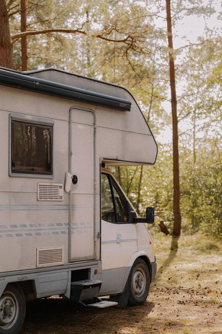 A white camper van with a large over-cab sleeping area is parked outdoors among tall trees with green foliage. The side of the vehicle features several small vents and a window, with the door closed. The van is situated on a dirt and grass surface, and the surrounding environment suggests a rural or woodland setting, consistent with outdoor home relocations or furniture transport preparations. The scene is lit with natural daylight, emphasizing the vehicle's exterior and the natural background. Man with Van Woodlands provides removals and packing services that may involve loading or unloading furniture and boxes, matching the context of vehicle loading process for house moves or relocation projects.