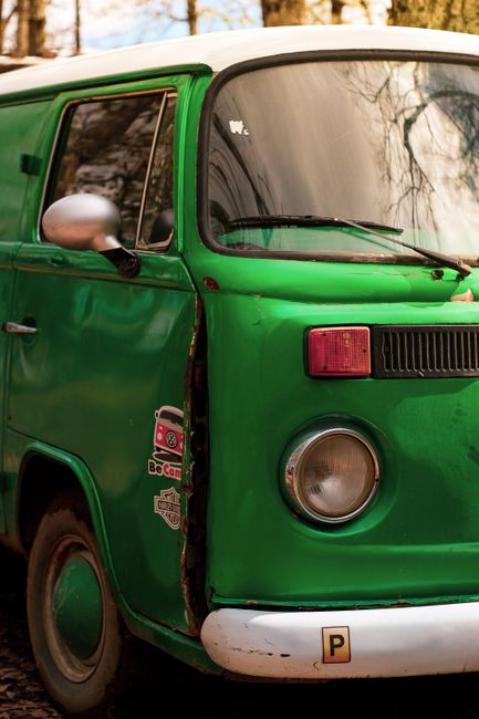 Close-up view of the front left side of a vintage green van parked outdoors, showing the passenger door and window, a side mirror, and a partial view of the front headlight and bumper. The van's surface has minor scratches and a sticker with the BeCoded logo is visible near the door. The vehicle is set against a blurred background of trees and natural surroundings, indicating an outdoor setting. This scene relates to home relocation and furniture transport, as vehicles like this are often used in moving services provided by Man with Van Woodlands, especially during the loading process of household items and packing materials for a house move.
