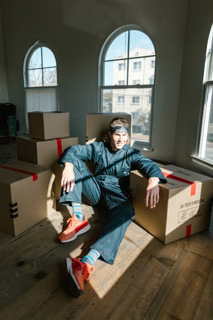 A young man with long hair, wearing a blue jumpsuit, a patterned headband, and bright orange sneakers, sits on a wooden floor inside a well-lit room with large arched windows showing a building and a tree outside. He is surrounded by several cardboard boxes, some sealed with red and black tape, and positioned in a way that suggests they are ready for packing or transport as part of a home relocation process. The man appears to be resting during a packing or loading phase, with a relaxed posture, one arm draped over a box and the other resting on his knee, embodying the logistics involved in furniture transport and moving services. The scene captures the interior environment, emphasizing the natural light coming through the windows, which highlights the boxes and the individual's work attire, typical of a professional removals service like Man with Van Woodlands involved in house removals and packing and moving operations.