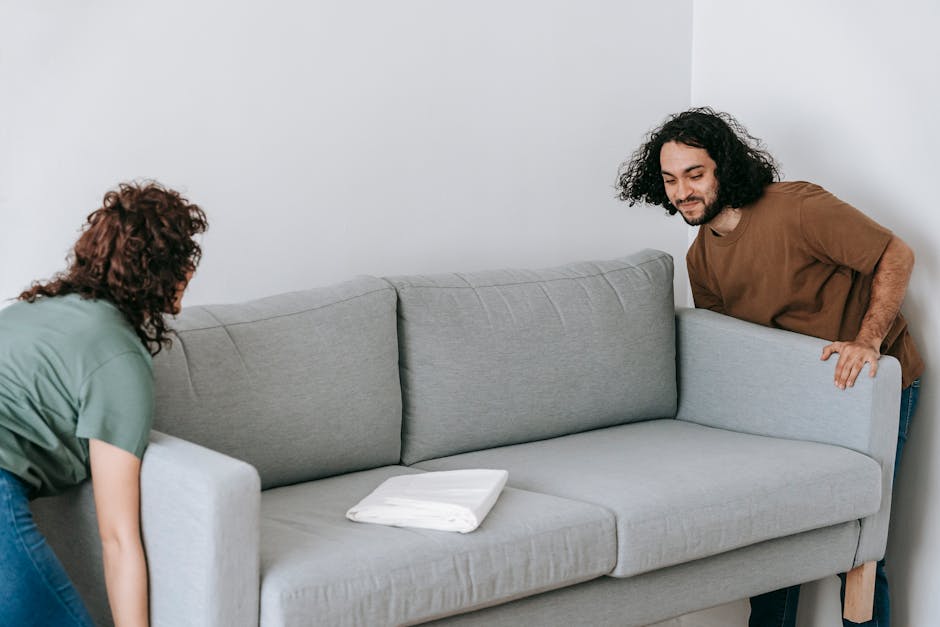 A man with shoulder-length curly hair and a brown T-shirt is seen gently lifting the armrest of a light grey fabric sofa inside a room with white walls, preparing for a home relocation. Beside him, a woman with curly brown hair in a green top is inspecting or assisting with the movement, her hands resting on the sofa cushion. On the sofa's seat, there is a folded white blanket or padding material, possibly used for protection during furniture transport. The room appears well-lit with natural or artificial light, and the sofa is positioned close to the white wall, indicating a typical setup in a household preparing for furniture packing and moving. This scene reflects the careful handling involved in furniture transport as part of a professional removal service, such as those offered by Man with Van Woodlands, emphasizing the logistics of moving bulky items like sofas through narrow spaces, consistent with house removal processes.