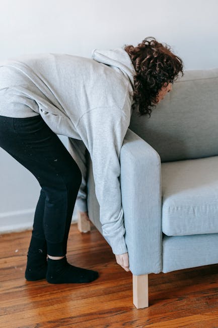 A person with curly brown hair wearing a light grey hoodie, black pants, and socks, is bending over and holding the edge of a grey fabric-upholstered sofa with wooden legs, positioned close to a white wall and wooden flooring. They appear to be preparing to move or adjust the furniture as part of a home relocation or furniture transport process. This scene takes place indoors, near a doorway or walking space, suggesting the initial stage of loading or moving furniture within a house. The image, representing packing and moving activities, highlights the careful handling of household items during a house removal, and may be associated with professional removals services such as those offered by Man with Van Woodlands, helpful in furniture transport and logistics when clearing or reorganising part of a property.