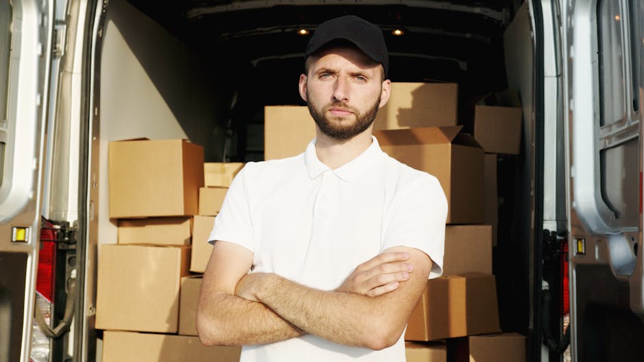 A young man with a serious expression, wearing a black cap and a white polo shirt, stands with his arms crossed inside the open back of a moving van. The van's interior is filled with multiple cardboa