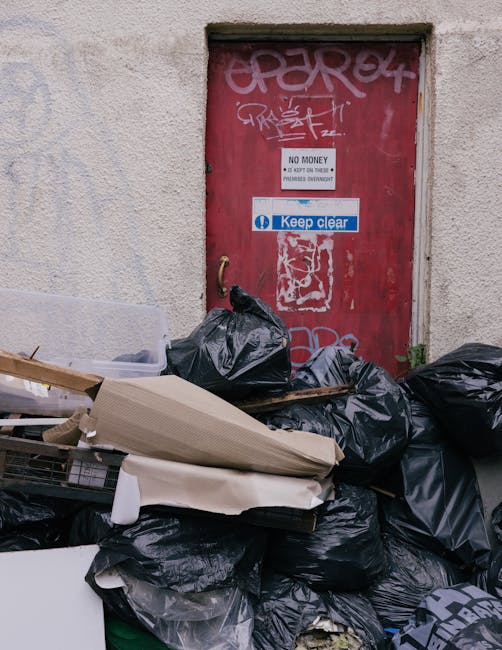 A red metal door with graffiti tags and various stickers, including a 'No Money' notice and a 'Keep Clear' sign, set into a beige concrete wall. In front of the door, there is a large pile of black rubbish bags, some packed tightly while others appear partially open, revealing contents like cardboard boxes and plastic packaging materials. There is a piece of cardboard leaning against the bags and a small white plastic tray resting on top. The scene is outdoors, on a paved area adjacent to the building, with natural daylight illuminating the clutter. This image captures the initial stages of home relocation or clearance, involving the collection of waste and packaging materials before transport, illustrating the importance of proper sorting in house removals. Man with Van Woodlands, a professional removals service, often assists with such logistics, ensuring that waste disposal and furniture transport are managed efficiently during moving processes.