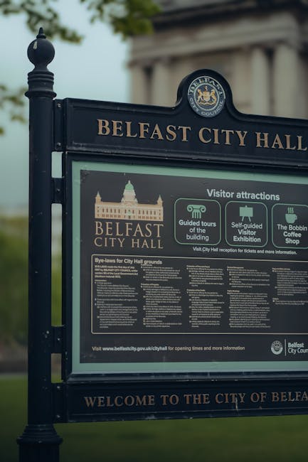 A black metal information board with the heading 'BELFAST CITY HALL' in gold lettering and the Belfast City Hall emblem at the top. The board displays details about visitor attractions, guided tours, self-guided visitor exhibitions, and the Bobbin Coffee Shop, with icons representing each feature. Below these, there are bye-laws for the City Hall grounds in fine print. The board is located outdoors on a paved area with a blurred background of green trees and cloudy sky, and it is supported by decorative black metal posts. This setting is typical for a tourist information display near a historic building, and the content supports the context of local attraction guidance relevant to house removals or relocation planning in Belfast, which aligns with [PAGE_TITLE] and [DOMAIN].