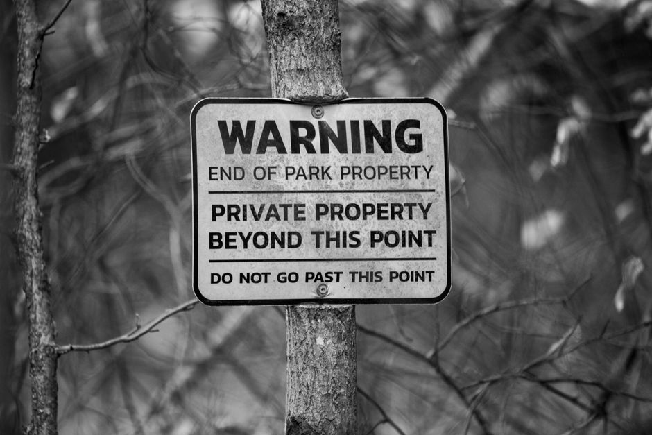 A black and white photograph of a warning sign attached to a tree trunk in an outdoor setting. The sign reads 'WARNING END OF PARK PROPERTY PRIVATE PROPERTY BEYOND THIS POINT DO NOT GO PAST THIS POINT' in bold uppercase letters, indicating restricted access. The background features blurred foliage and tree branches, suggesting a wooded area. This image highlights the importance of respecting property boundaries, relevant for house removals and furniture transport services provided by Man with Van Woodlands, especially during packing and loading processes in outdoor environments.